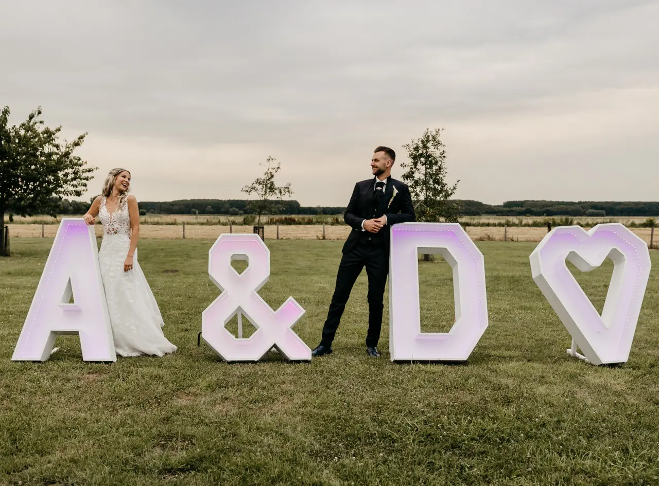 Een bruidspaar bij de verlichte letters A en D met een hart, op een grasveld bij valavond.