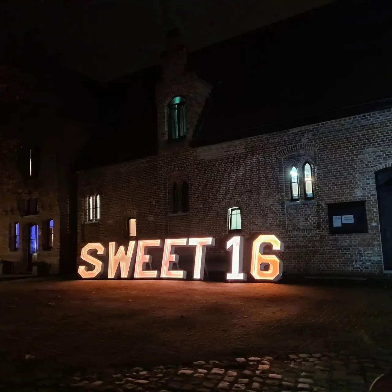 SWEET 16 in verlichte letters tegen een bakstenen gevel.