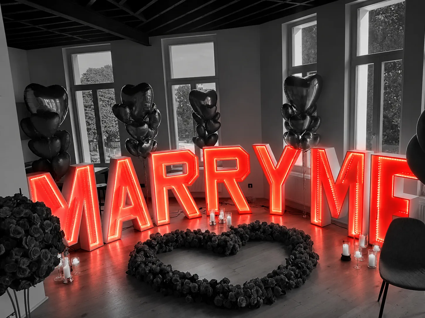 Een huwelijksaanzoek: de letters MARRY ME achter een hart van rozen, met rode hartballonnen.