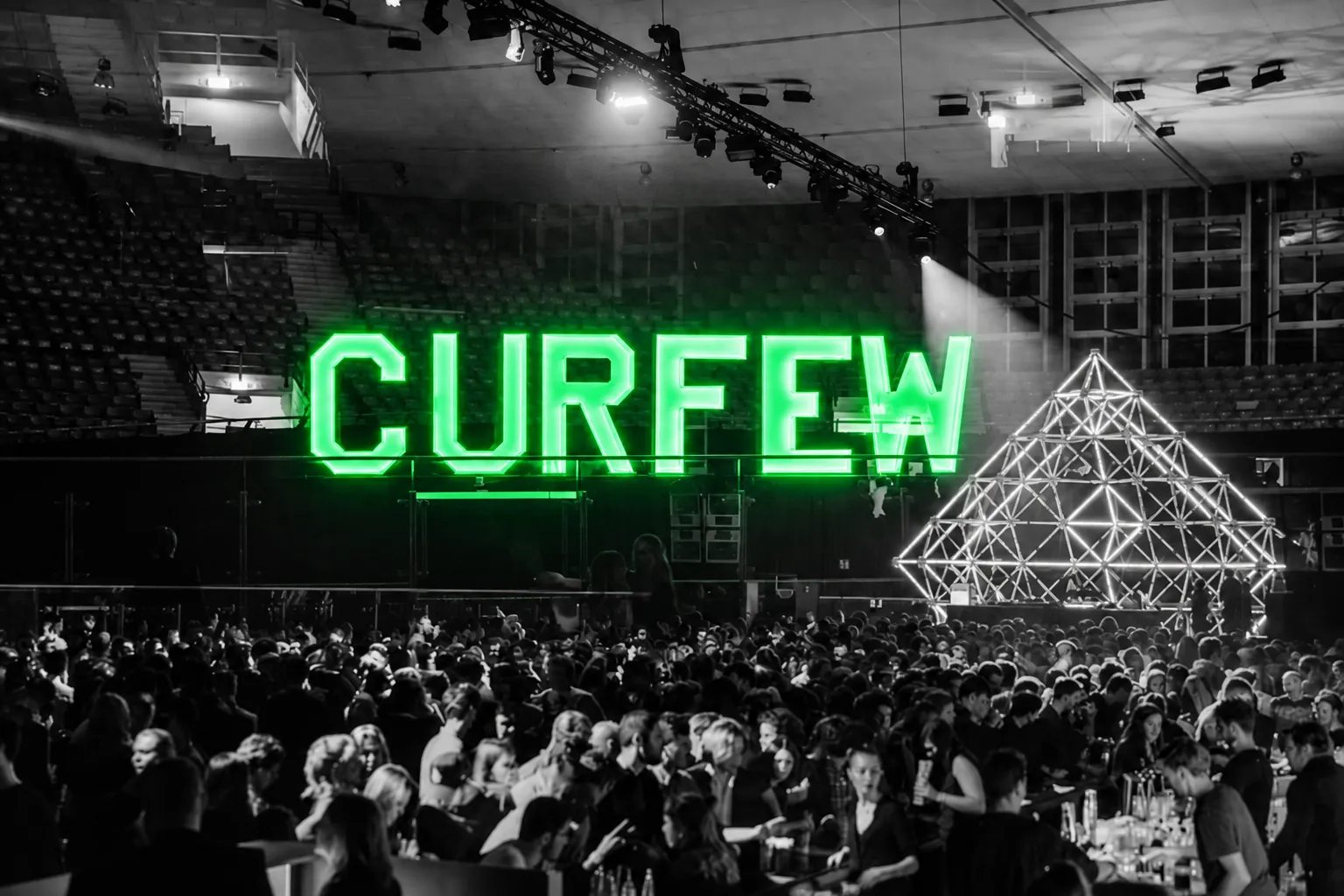 De groene letters CURFEW boven een volle feestzaal met podiumlicht.